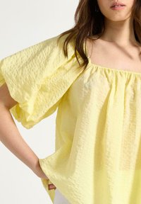 Gul, texturerad blus med puffade korta ärmar, elastisk halsringning och lös passform. Tyget verkar lätt och andningsbart.