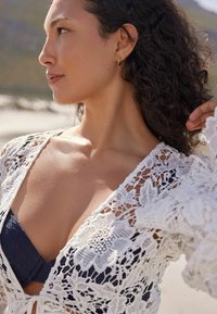 Biała koronkowa narzutka z kwiatowymi wzorami i otwartym splotem, nakładana na granatowy bikini. Kręcone włosy oprawiające twarz, naturalny odcień skóry.