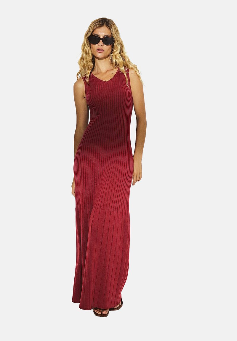 Abito maxidress rosso, a coste, senza maniche, con scollatura a V, corpetto aderente e gonna svasata. Indossato con occhiali da sole scuri e sandali aperti.