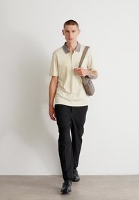 Beige polo skjorte med en gråmønstret krage, parret med svarte bukser og svarte sko, mens han bærer en grå skulderveske.