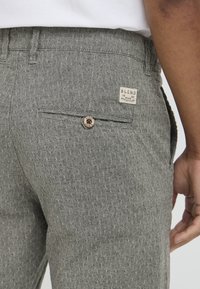 Pantaloni grigi con texture e un motivo sottile, dotati di una tasca posteriore, un'etichetta con marchio e un bottone marrone sulla chiusura della tasca.