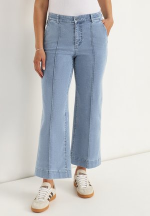 Person iført lyseblå vidde jeans med lodrette sømme og hvide sneakers med sorte striber.