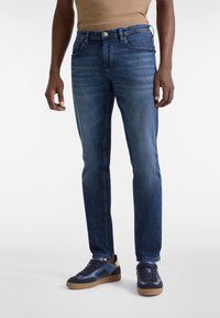 Hombre vistiendo jeans de denim azul ajustados y zapatillas de deporte azul marino con suelas de goma, de pie contra un fondo simple.