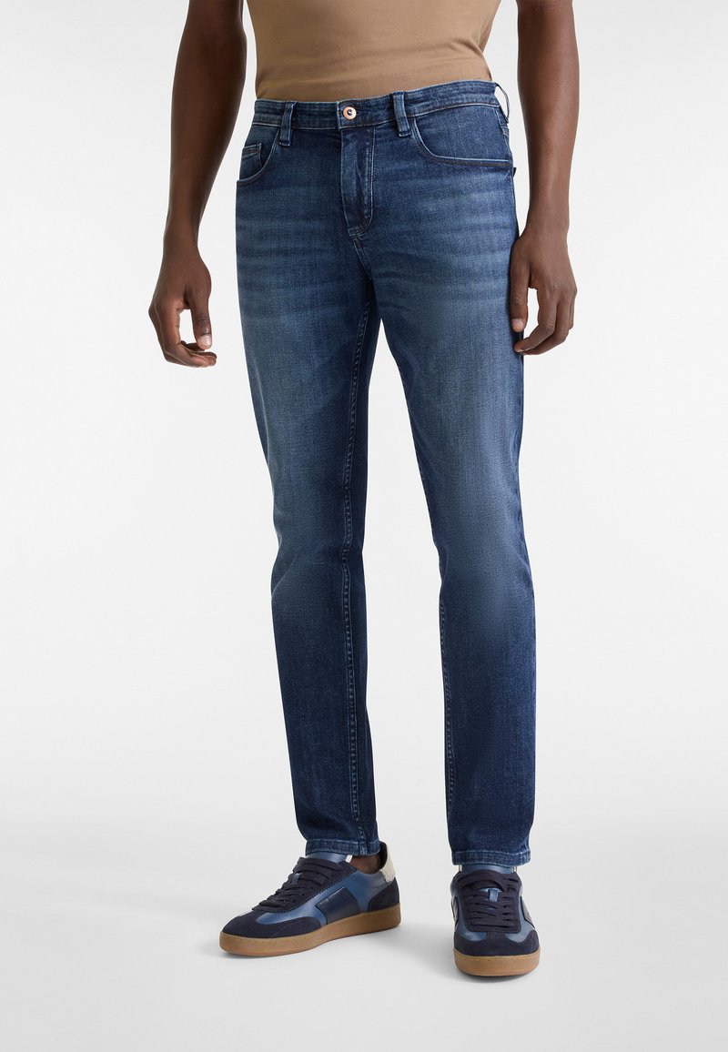 Hombre vistiendo jeans de denim azul ajustados y zapatillas de deporte azul marino con suelas de goma, de pie contra un fondo simple.
