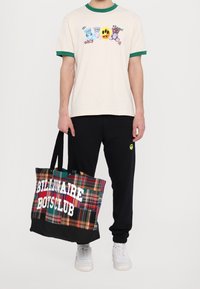 Osoba ubrana w beżowy t-shirt z rysunkami niedźwiedzi, czarne dresy z żółtym logo uśmiechu, trzymająca wielokolorową torbę w kratę z białym napisem.