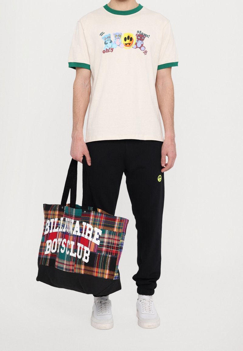 Osoba ubrana w beżowy t-shirt z rysunkami niedźwiedzi, czarne dresy z żółtym logo uśmiechu, trzymająca wielokolorową torbę w kratę z białym napisem.