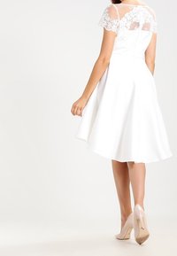 Bílé šaty s krajkovými cap rukávy a přiléhavým korzetem, vycházející sukně ve tvaru písmene A a částečně průhledný zadní díl. Neutrální vysoké podpatky.