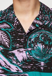 Chemise à manches courtes avec un fond noir orné de motifs tropicaux multicolores en turquoise et lavande, incluant des feuilles et des oiseaux.