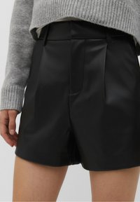 Pantalones cortos negros de cuero sintético plisados con un ajuste entallado. Presentan una textura suave y una cintura. Combinados con un suéter de punto gris.
