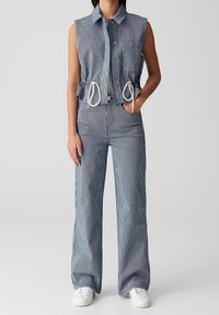 Gilet sans manches à col avec boutons sur le devant et taille ajustée par un cordon, associé à un pantalon large à fines rayures assorti et des baskets blanches.