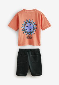 Oranje katoenen T-shirt met een blauwe zonafdruk op de achterkant, gecombineerd met zwarte shorts met vier zakken en witte stiksels.