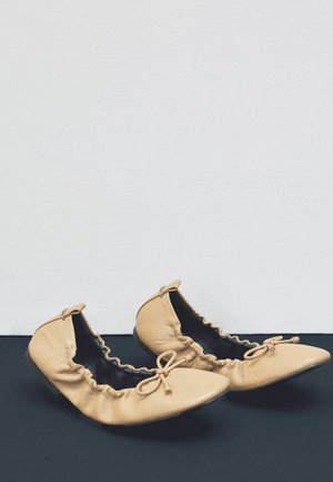 Nude színű bőr balettcipők reszelhető szélekkel, hegyes orral és díszes masnikkal az elején, fekete felületen, fehér háttér előtt pihennek.