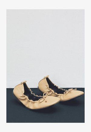 Goli čevlji za balet iz usnja v kožni barvi z nabranimi robovi, koničastimi prsti in okrasnimi mašnjami spredaj, počivajo na črni površini na belem ozadju.