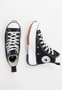 Sneaker alte in tela nera con lacci bianchi, punta in gomma e suola testurizzata a contrasto in marrone e nero. Logo Chuck Taylor visibile.