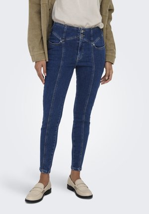 Jeans ajustados de mezclilla azul con cintura alta, cierre de doble botón, detalles de costura sutiles y una textura suave. Llevados con mocasines beiges.
