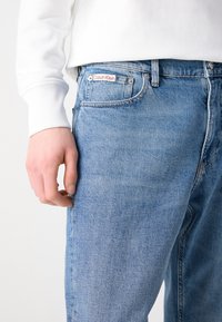 Lyseblå denimjeans med lige snit, med et rødt og hvidt "Calvin Klein" logo-patch på taljen og synlige stikninger.
