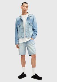 Ljusblå denimjacka med grå huva, vit t-shirt, ljusblå shorts och svarta slip-on skor med vita strumpor, ståendes på en grå bakgrund.
