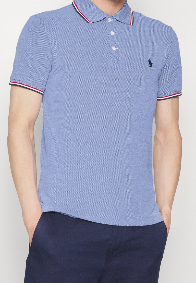 Homme portant un polo bleu clair avec un liseré rouge et blanc au col et aux manches, pantalon bleu marine, mains dans les poches, debout devant un fond blanc.