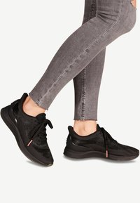 Chaussures de sport noires avec une tige en mesh texturé, un design à lacets et un accent orange contrastant sur le talon, associées à un jean gris.