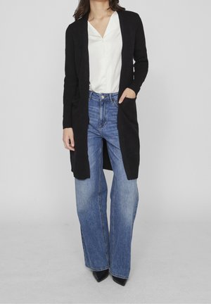 Czarny długi kardigan z prążkowanymi mankietami i kieszeniami, noszony na białej bluzce i niebieskich spodniacjach z wysokim stanem i szerokimi nogawkami. Gładka tekstura, casualowy design.