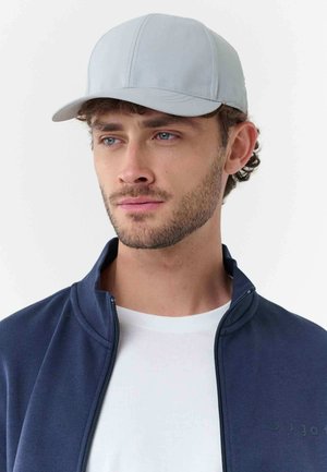 Young man with light facial hair wearing a light gray cap, white shirt, and navy blue zip-up jacket, looking slightly to the side.
