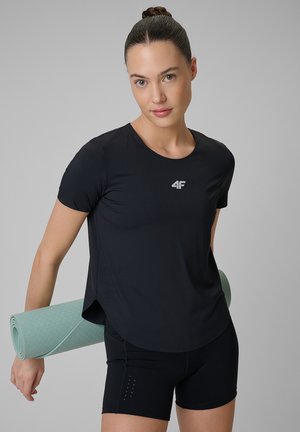 Kobieta w czarnym stroju sportowym trzyma zwiniętą jasnoniebieską matę do jogi na jednolitym szarym tle.