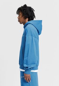 Modrá mikina s kapucí vyrobená z měkké bavlny, s klokaní kapsou, žebrovanými manžetami a odpovídajícím outfitem. Obsahuje malé logo na hrudi.