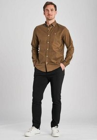 Cămașă maro cu nasturi, guler, nasturi albi și un logo subtil, asortată cu pantaloni negri și adidași albi. Design și croială simple.