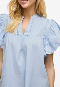 Gaiši zils blūze ar piepūstām piedurknēm, V veida kakla izgriezumu un vertikāliem krokiem. Iekļautas smalkas mežģīņu detaļas gar kakla izgriezumu. Mīksta auduma tekstūra.