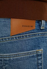Blå denimjeansficka med en brun läderlapp märkta "CHASIN'" under en brun ribbad tröjkant.
