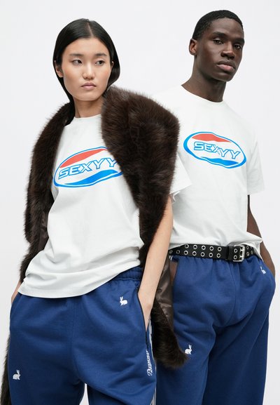 Vita t-shirts med texten "SEXYYY" i blått och rött, kombinerade med blå mjukisbyxor med kaninlogotyper och ett svart nitat bälte.