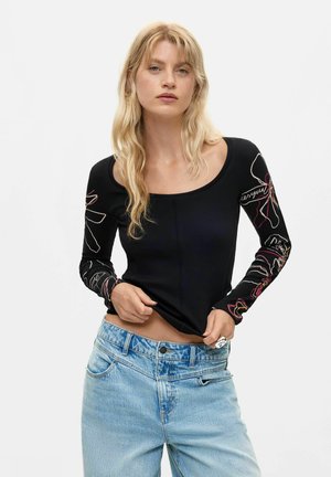 Mujer rubia con una camiseta negra de manga larga con diseños florales en línea y vaqueros azul claro, levantando ligeramente el dobladillo de su camisa.