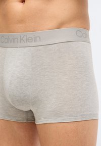 Κοντινό πλάνο ενός ατόμου που φοράει ανοιχτόγριες αντρικές μποξεράκια Calvin Klein, παρουσιάζοντας τη λαστιχένια μέση και την περιοχή του άνω μηρού.