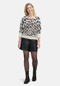 Usmievajúca sa žena v béžovom svetri s leopardím vzorom, čiernych kožených šortkách, čiernych priesvitných pančuchách a čiernych členkových topánkach stojaca na bielom pozadí.