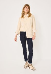 Camicetta beige con maniche arrotolate, abbinata a jeans skinny scuri e sneaker beige con dettagli marroni. Tessuto morbido e vestibilità classica.