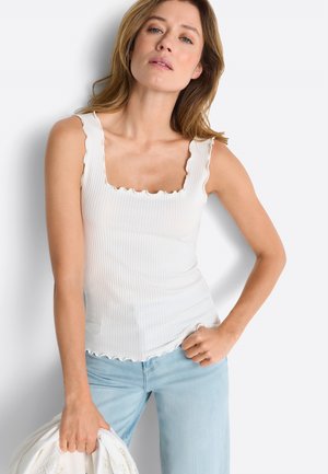 Femme aux cheveux ondulés portant un débardeur côtelé blanc avec des bords festonnés et un jean bleu clair, tenant une veste blanche dans sa main gauche.