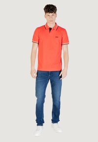 Polo rosso con colletto nero e bordi bianchi, abbinato a jeans blu e sneakers bianche. Il modello si trova in un ambiente neutro.