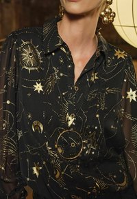 Blusa nera con stampa celestiale dorata, caratterizzata da stelle e costellazioni, con bottoni frontali e una consistenza morbida. Include una collana circolare dorata.