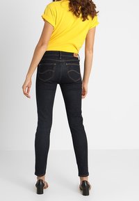 Jeans skinny azul oscuro con costuras finas, de cintura alta y un parche con logo en la parte posterior. Combinados con una blusa amarilla de mangas enrolladas y zapatos de tacón.