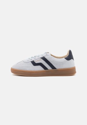 Weiße Wildleder-Sneaker mit navyblauen Akzenten, ausgestattet mit einer Gummisohle, runder Zehenpartie und hellblauen Schnürsenkeln. Das GANT-Logo ist an der Seite angebracht.