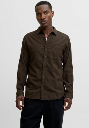 Young man wearing a dark brown button-up shirt with a chest pocket and black pants, standing against a plain light background.