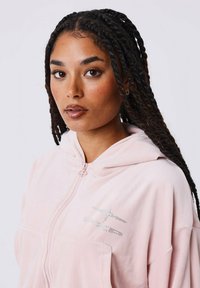 Felpa rosa con zip realizzata in tessuto morbido. Caratterizzata da un cappuccio con cordino e un logo in strass argentati sul petto. Tessuto liscio e vestibilità casual.