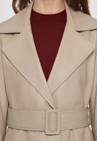 Bežinė dirbtinės odos trenčkotė su plačiomis apykaklėmis, diržu talijoje ir matomomis siūlėmis, uždėta ant bordo spalvos viršaus.