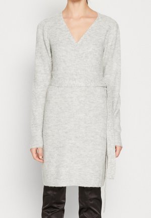 Kobieta ubrana w jasnoszarą dzianinową sukienkę kopertową z długimi rękawami i dekoltem w serek, w połączeniu z czarnymi spodniami.