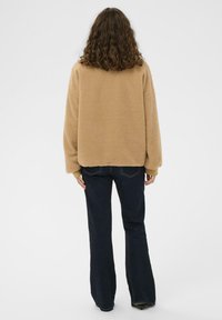 Beige fliisjakker, millel on lõdvestatud sobivus ja madalad õlavarred, koos tumedate laienevate teksadega. Kanga välimus on pehme ja tekstuurne.