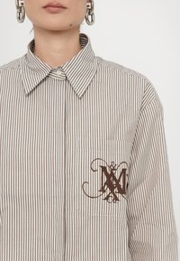 Striped shirt in beige and brown with a button collar, featuring a left chest pocket and a decorative brown logo embroidery.