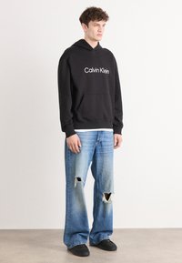 Juoda gobtuvinė palaidinė su "Calvin Klein" logotipu, derinama su šviesiai mėlynomis nusidėvėjusiomis plačiomis kelnėmis, turinčiomis suplėšymų detalių. Juodi batai.
