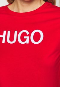 Červené bavlněné tričko s velkým bílým logem "HUGO". Design zahrnuje klasický kulatý výstřih a krátké rukávy. Měkká textura.
