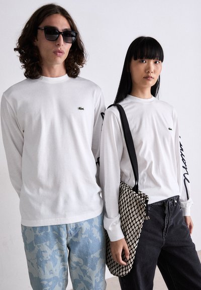 Deux personnes portent des t-shirts blancs à manches longues avec un logo vert. La première est associée à un jean à motifs bleu clair ; la seconde tient un sac en maille noir et blanc.