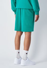 Grüne Baumwollshorts mit einem elastischen Bund, kombiniert mit weißen Socken und Turnschuhen. Der Stoff wirkt weich und hat eine glatte Textur.